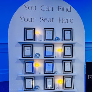 May include: A white seating chart with the text "You Can Find Your Seat Here". It features framed cards with names and numbers, decorative candles, and disco balls. The background is blue.