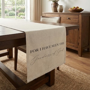 May include: A beige linen table runner with the text "FOR I HAVE SEEN THE Goodness of God" in black script. The runner is draped over a dark brown wooden table. A wooden cabinet and chair are in the background.