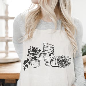May include: A cream-colored apron featuring a black and white illustration of potted plants. The design includes a stack of pots, a pot with trailing vines, and a pot with tall grass.