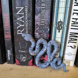 Peut inclure: Une figurine de serpent bleu clair moucheté avec des accents dorés, posée devant une rangée de livres. Le serpent est enroulé et détaillé, ajoutant une touche décorative à une bibliothèque.