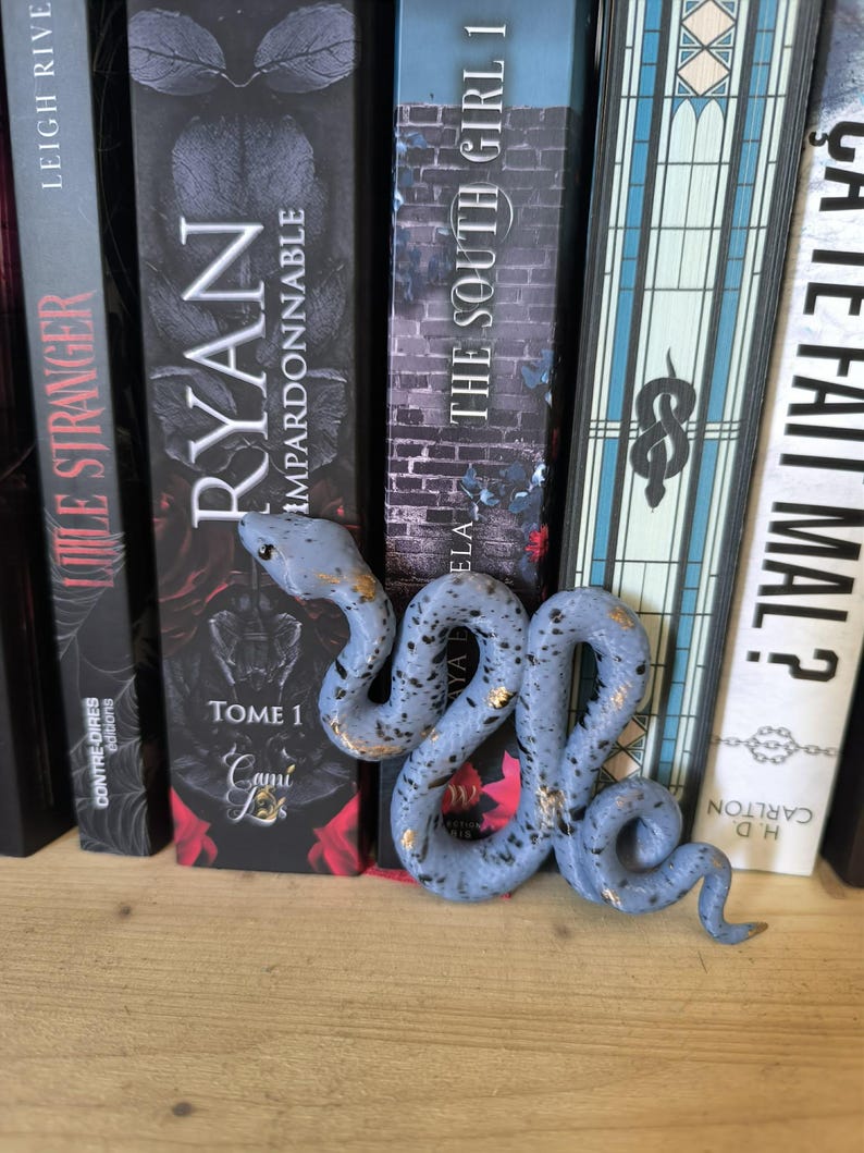 Puede incluir: Una figura de serpiente azul claro moteada con detalles dorados, colocada frente a una fila de libros. La serpiente est&aacute; enrollada y detallada, agregando un toque decorativo a una estanter&iacute;a.