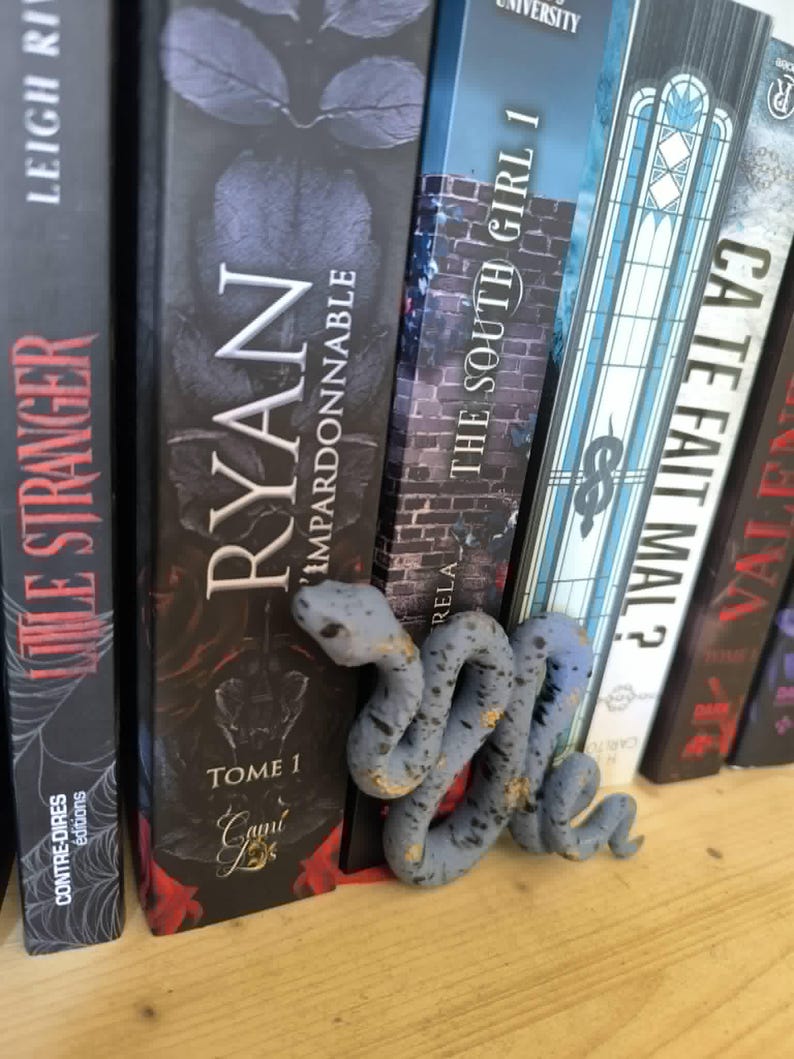 Puede incluir: Un marcap&aacute;ginas en forma de serpiente gris y dorada descansa entre una fila de libros. El marcap&aacute;ginas est&aacute; enrollado y detallado, con un aspecto texturizado. Los libros tienen cubiertas oscuras con t&iacute;tulos como "RYAN" y "THE SOUTH GIRL 1".