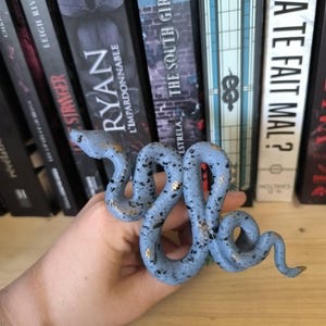 Puede incluir: Una figura de serpiente de cer&aacute;mica azul claro con motas negras y detalles dorados. La serpiente est&aacute; enrollada y tiene una superficie detallada y texturizada. La figura se exhibe frente a una estanter&iacute;a.