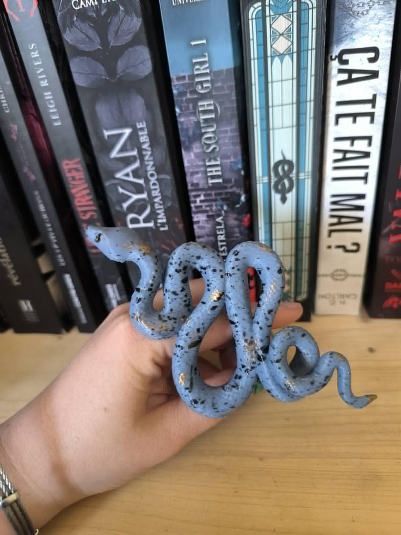 Puede incluir: Una figura de serpiente de cer&aacute;mica azul claro con motas negras y detalles dorados. La serpiente est&aacute; enrollada y tiene una superficie detallada y texturizada. La figura se exhibe frente a una estanter&iacute;a.