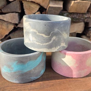 May include: Three small, cylindrical concrete planters in shades of gray, blue, and pink. The planters have a marbled effect and are stacked on top of each other. The background features a stack of firewood.