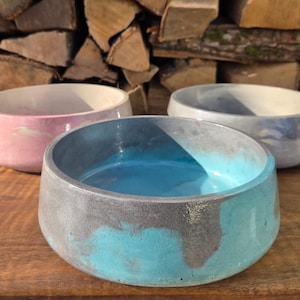 May include: Three handcrafted concrete bowls in shades of pink, blue, and gray. The bowls are round with a smooth finish and sit on a wooden surface. The background features stacked firewood.