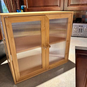 May include: A light brown bamboo cabinet with two doors and clear, textured glass panels. The cabinet has two wooden shelves inside and wooden knobs. The cabinet is designed for storage and organization.