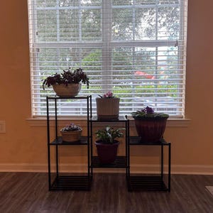 May include: A black metal plant stand with three tiers, displaying several potted plants. The plants feature green foliage and flowers, with pots in brown, beige, and purple hues. The stand is placed in front of a window with slatted blinds.
