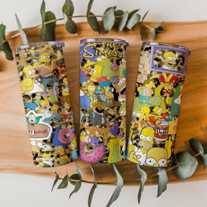 May include: Three stainless steel tumblers featuring a colorful collage of The Simpsons characters. The designs include Homer, Bart, Lisa, Marge, and other characters, set against a leopard print background. The tumblers are displayed on a wooden surface.