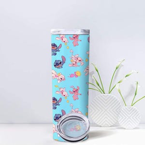 May include: A cylindrical tumbler with a light blue background featuring a repeating pattern of blue and pink cartoon characters. The tumbler has a clear plastic lid with a sliding closure, and the design includes small yellow heart accents.