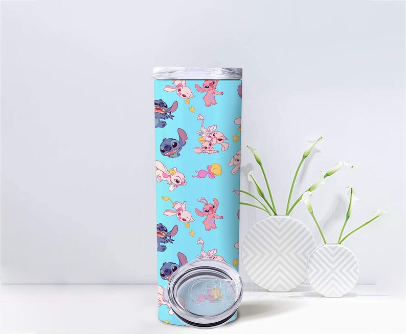 May include: A cylindrical tumbler with a light blue background featuring a repeating pattern of blue and pink cartoon characters. The tumbler has a clear plastic lid with a sliding closure, and the design includes small yellow heart accents.