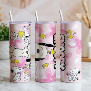May include: Three cylindrical tumblers with white straws, featuring a light pink background with a repeating pattern of Snoopy and Woodstock characters alongside pink flower petals. The text Snoopy is printed vertically in black on the tumblers.