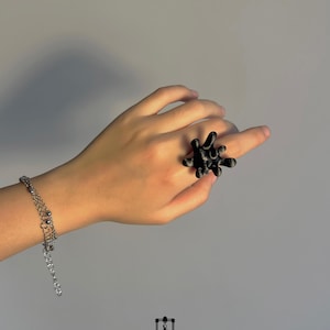 Könnte beinhalten: Ein schwarzer, abstrakter Ring mit einem einzigartigen, stacheligen Design wird an einer Hand getragen. Ein silbernes Armband mit mehreren Ketten ist ebenfalls sichtbar. Der Ring ist der Blickfang und zeigt eine moderne, künstlerische Ästhetik. Der Text "LUMO Sculpture 3D Studio" ist enthalten.