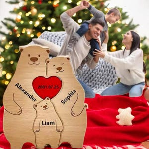 Peut inclure: Puzzle en bois représentant une famille d'ours avec les noms "Alexander", "Sophia" et "Liam" gravés. Un cœur rouge central affiche la date "2001.07.25". L'ensemble est posé sur une couverture rouge, avec un sapin de Noël en arrière-plan.
