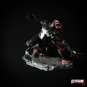 Puede incluir: Figura de Venom en blanco y negro en una pose din&aacute;mica, con detalles rojos en la boca y las manos. La figura lleva zapatillas rojas y blancas y est&aacute; sobre una base de piedra gris. Tiene un emblema de ara&ntilde;a blanca en el pecho.