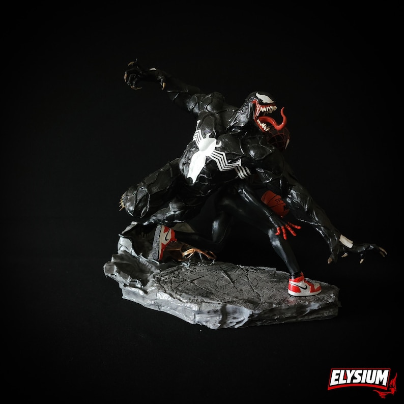 Puede incluir: Figura de Venom en blanco y negro en una pose din&aacute;mica, con detalles rojos en la boca y las manos. La figura lleva zapatillas rojas y blancas y est&aacute; sobre una base de piedra gris. Tiene un emblema de ara&ntilde;a blanca en el pecho.