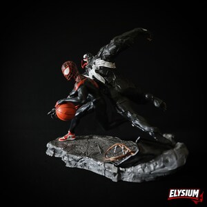 Puede incluir: Una figura din&aacute;mica con Miles Morales Spider-Man botando un bal&oacute;n de baloncesto, con Venom saltando detr&aacute;s de &eacute;l. Las figuras son predominantemente negras y rojas, sobre una base gris texturizada. El bal&oacute;n es naranja y las zapatillas son rojas y blancas.