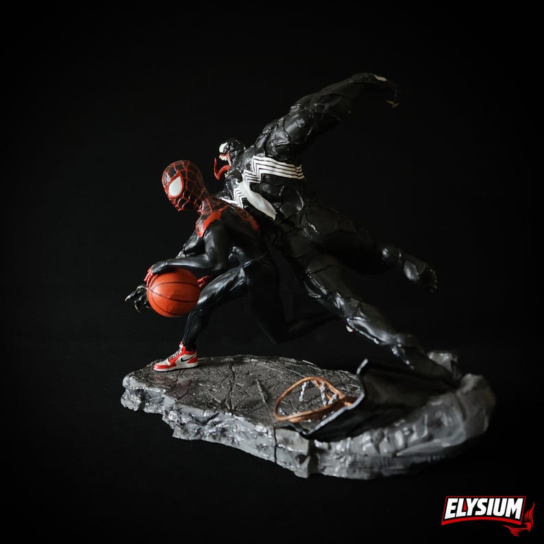 Puede incluir: Una figura din&aacute;mica con Miles Morales Spider-Man botando un bal&oacute;n de baloncesto, con Venom saltando detr&aacute;s de &eacute;l. Las figuras son predominantemente negras y rojas, sobre una base gris texturizada. El bal&oacute;n es naranja y las zapatillas son rojas y blancas.