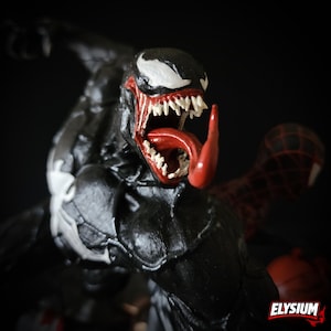 Puede incluir: Figura de acci&oacute;n negra de Venom con un emblema de ara&ntilde;a blanco en el pecho y la cabeza. La figura tiene una lengua roja y afilados dientes blancos. Una figura de Spider-Man roja y negra est&aacute; en el fondo. La palabra "ELYSIUM" est&aacute; en la esquina inferior derecha.