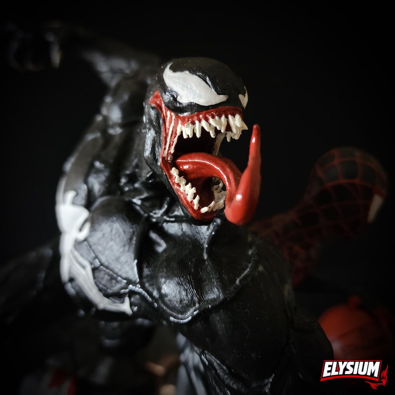 Puede incluir: Figura de acci&oacute;n negra de Venom con un emblema de ara&ntilde;a blanco en el pecho y la cabeza. La figura tiene una lengua roja y afilados dientes blancos. Una figura de Spider-Man roja y negra est&aacute; en el fondo. La palabra "ELYSIUM" est&aacute; en la esquina inferior derecha.