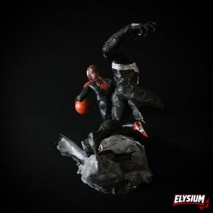 Puede incluir: Figura detallada con dos personajes de c&oacute;mic. Una figura viste un traje negro con detalles blancos, y la otra un traje rojo y negro, sosteniendo un bal&oacute;n de baloncesto naranja. Las figuras posan sobre una base de roca gris sobre un fondo negro.