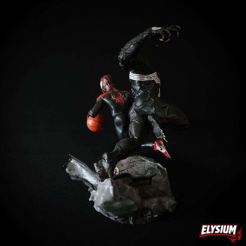 Puede incluir: Figura detallada con dos personajes de c&oacute;mic. Una figura viste un traje negro con detalles blancos, y la otra un traje rojo y negro, sosteniendo un bal&oacute;n de baloncesto naranja. Las figuras posan sobre una base de roca gris sobre un fondo negro.
