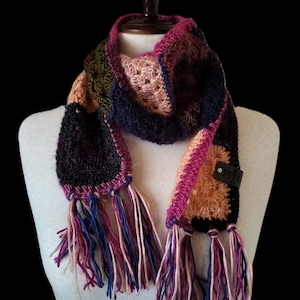 May include: A handmade crochet scarf in a variety of colors, including black, pink, orange, blue, and green. The scarf has a fringed edge with multiple strands of yarn. The scarf is displayed on a mannequin.