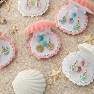 Può includere: Un assortimento di scatole portagioie a forma di conchiglia, ciascuna contenente un paio di orecchini. Gli orecchini presentano vari design, tra cui floreali, a farfalla e geometrici, in tonalità oro, rosa e blu. Le scatole sono disposte su una superficie sabbiosa con stelle marine.