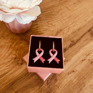 Peut inclure: Une paire de boucles d'oreilles créoles dorées avec des breloques en forme de ruban rose dans deux nuances de rose. Les boucles d'oreilles sont présentées dans une boîte carrée rose avec un insert en velours noir pour maintenir les bijoux.