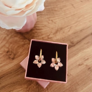 Könnte beinhalten: Ein Paar goldfarbene Creolen mit rosa blütenförmigen Anhängern, präsentiert in einer rosa Geschenkbox. Die Ohrringe liegen auf einer Holzoberfläche, mit einer rosa Blume im Hintergrund.