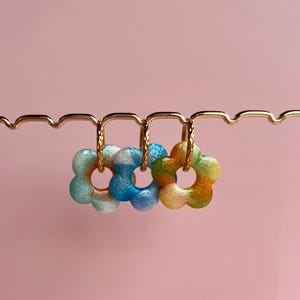 Op de afbeelding: Drie bloemvormige oorbellen in blauwe, groene en gele tinten hangen aan een goudkleurige displaybalk. De oorbellen hebben een gemarmerd uiterlijk tegen een zachtroze achtergrond. Een uniek accessoire.