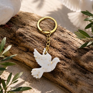 Könnte beinhalten: Ein weißer Schlüsselanhänger in Taubenform mit eingearbeiteten goldfarbenen Sprenkeln. Der Anhänger ist mit einer kurzen Kette an einem goldfarbenen Metallring befestigt und bildet ein dekoratives Accessoire mit glatter, glänzender Oberfläche.