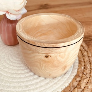 Peut inclure: Un bol en bois de couleur claire avec une bande noire. Le bol est rond avec une large ouverture et repose sur un tapis tissé blanc et beige. Un vase rose avec une fleur blanche est en arrière-plan.