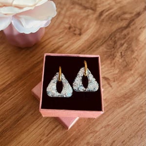 Peut inclure: Une paire de boucles d'oreilles triangulaires avec une finition texturée blanc irisé et argenté, dotées d'une découpe centrale. Chaque boucle est fixée à un petit anneau en métal doré poli. Les boucles sont présentées dans une boîte cadeau rose.