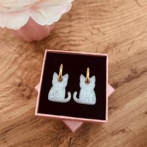 Puede incluir: Pendientes con forma de gato con aros dorados, presentados en una caja de regalo rosa con interior negro. Los pendientes son de color azul claro y blanco, con un aspecto texturizado. La caja está sobre una superficie de madera, con una flor rosa en el fondo.