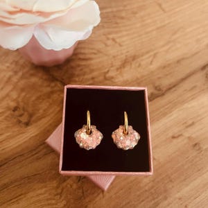 Op de afbeelding: Een paar oorbellen in de vorm van een zeeschelp met gouden ringen, gepresenteerd in een roze geschenkdoos. De oorbellen zijn lichtroze met glitteraccenten. Op de achtergrond is een roze bloem te zien.