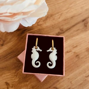 May include: A pair of earrings featuring white seahorse-shaped charms suspended from gold-toned hoop rings. The earrings are presented inside a small, square, pink-coloured gift box with a black velvet insert, showcasing the detailed seahorse design.