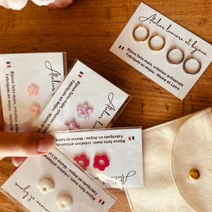 Könnte beinhalten: Mehrere Paare von Ohrsteckern und Creolen auf weißen Marken-Karten mit der Aufschrift Atelier lumière et bijoux. Die Ohrringe umfassen durchscheinende rosa Blumen, glitzernde Sterne, einfarbige magentafarbene Blumen, weiße strukturierte Kreise sowie goldene und silberne Creolen.