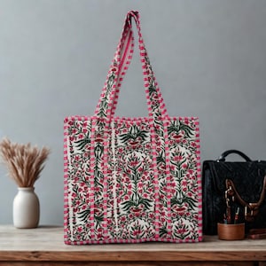 May include: A patterned tote bag featuring a floral design with red flowers, green leaves, and a white background. The bag has pink trim and handles. A vase and a black bag are in the background.