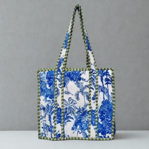 May include: A white tote bag with a blue tropical print depicting palm trees and animals. The bag has green and white striped trim and handles. The bag is designed for carrying items.