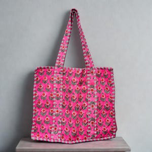 May include: A pink tote bag with a floral design. The bag has a repeating pattern of red flowers and green leaves on a pink background. The handles are made from the same patterned fabric. The bag is on a wooden surface.