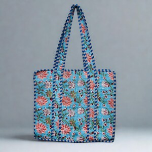 May include: A blue tote bag featuring a floral design in red, white, and green. The bag has blue and white striped handles and trim. It is constructed from quilted fabric.