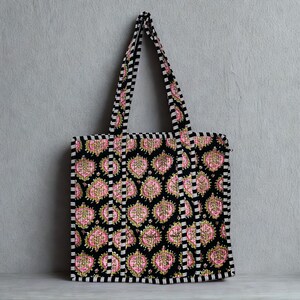 May include: A black tote bag featuring a repeating pink and gold floral design. The bag has black and white striped trim and handles. The tote bag is suspended against a grey backdrop.