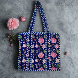 May include: A blue tote bag with a floral pattern of pink flowers and green leaves. The bag has blue and white striped handles and trim. The bag is made of fabric and is on a grey surface.