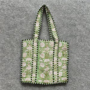 May include: A quilted tote bag with a floral pattern in shades of green and white. The bag has sturdy handles and a striped border. The bag is designed for carrying items.