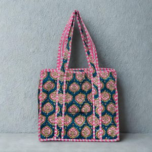 May include: A quilted tote bag featuring a teal background with a repeating pink and gold floral design. The bag has pink and white striped handles and trim. The bag is designed for carrying items.