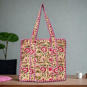 May include: A quilted tote bag with a floral pattern in shades of pink, yellow, and green. The bag has a rectangular shape with matching pink handles and trim. The bag is sitting on a wooden surface.