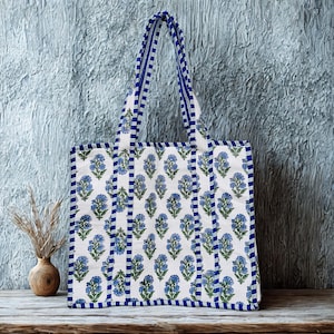May include: White tote bag featuring a blue floral design and blue striped edging. The bag has matching blue handles. The bag is made of a durable fabric and is suitable for carrying shopping or other items.
