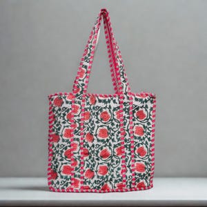 May include: A floral patterned tote bag. The bag has a white base with red flowers and green foliage. The edges and handles are trimmed with a bright pink fabric. The bag is designed for carrying items.