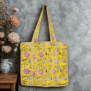 May include: Yellow tote bag featuring a floral design in pink, white, and black. The bag has long straps and a quilted texture. The background is a grey wall with a vase of flowers on a wooden table.
